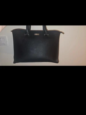 Handväska - Fin handväska från Don Donna.  Egentligen sparsamt använd, har de senaste cirka 8 åren endast legat i en garderob.  På insidan av handtagen (x2) har trådar släppt, var god se bilder. Det ska nog vara ett medföljande axelband till, men det vet jag tyvärr inte var det finns.  Ett fack med dragkedja på baksidan av väskan. Insidan har 3 fack, varav ett stängs med dragkedja. 2 små fack på insidan, ett "öppet" fack och ett med dragkedja. Alla 3 fack är enligt mitt tycke rymliga.