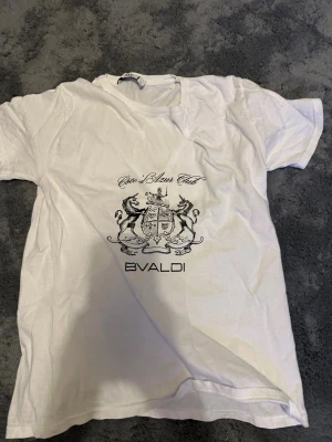 Vit t-shirt från Bvaldi med tryck - Vit t-shirt från Bvaldi med svart tryck framtill som visar ett stilrent vapensköldmotiv och texten 'Côte d’Azur Club'. Klassisk passform och rund halsringning, perfekt för en clean och stilren look.