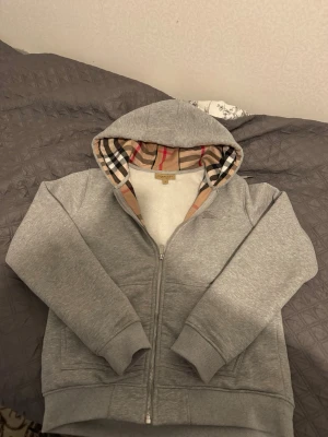 Grå zip hoodie från Burberry - Snygg grå zip hoodie från Burberry med klassiskt rutigt mönster i huvan. Hoodien har dragkedja framtill, två fickor och ribbade muddar vid ärmar och nederkant. Perfekt för en chill och stilren look.