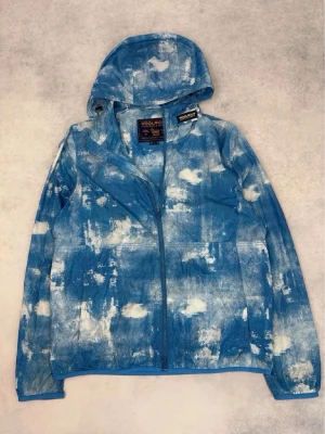 Blåvit vindjacka från Woolrich - Säljer en blåvit vindjacka från Woolrich med coolt batikmönster och hel dragkedja framtill. Jackan har huva och två sidofickor. Perfekt för dig som vill sticka ut med en unik och trendig look. Materialet känns lätt och smidigt.