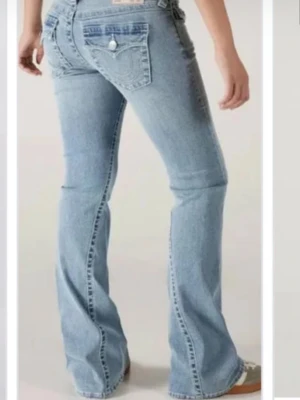 Ljusblå bootcut jeans från True Religion - Säljer ett par ljusblå bootcut jeans från True Religion med klassiska kontrastsömmar och ikoniska fickdetaljer bak. Jeansen har låg midja och utsvängda ben, perfekta för en avslappnad och trendig look.