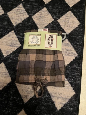 Rutiga flanellbyxor från Arctic Frog - Mysiga pyjamasbyxor i flanell från Arctic Frog. Byxorna är rutiga i beige, grått och svart, har dragsko i midjan och två framfickor. Tillverkade i 100% bomull för extra komfort, perfekta för chill hemma.ORDINARIE PRIS:200kr