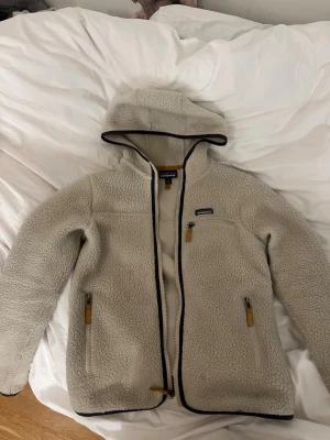Beige teddyjacka från Patagonia - Mysig beige teddyjacka från Patagonia med huva och dragkedja. Jackan har tre fickor.Materialet är fluffigt och ger en avslappnad look.