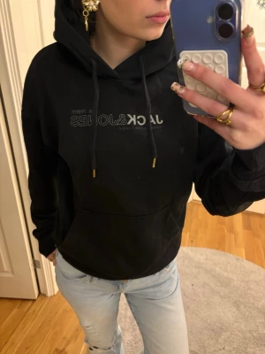 Svart hoodie från Jack & Jones - Säljer en svart hoodie från Jack & Jones med tryck på bröstet och ryggen. Hoodien har en stor ficka framtill, justerbar huva med snören och ribbade muddar vid ärmar och midja. Perfekt för en chill och avslappnad stil.