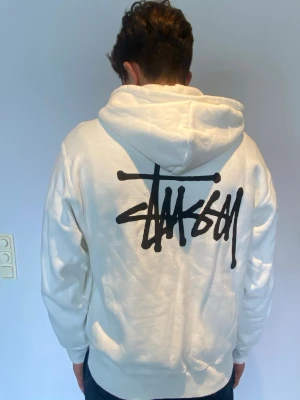 Vit zip hoodie från Stüssy - Säljer en vit hoodie från Stüssy med dragkedja, stor huva och två fickor fram. Stüssy-logga i svart tryck på bröstet och stort tryck på ryggen. Tillverkad i mjukt bomullsmaterial, perfekt för en avslappnad streetwear-look. Äkta och köpt från Stüssy’s hemsida. Pris kan förhandlas 