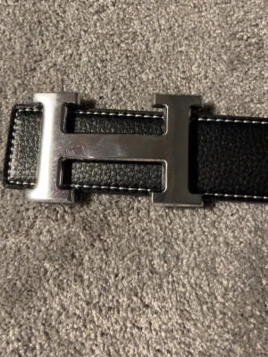 Svart skärp med H-spänne i silver - Stilrent svart skärp med ett stort H-format spänne i silverfärg. Skärpet har vita sömmar längs kanterna och är gjort i slätt skinn. Perfekt accessoar för att lyfta din outfit. Ta insta box helst som frakt 