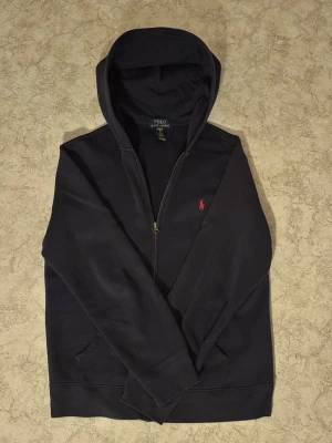 Mörkblå hoodie från Polo Ralph Lauren - Snygg mörkblå hoodie från Polo Ralph Lauren med dragkedja och klassisk röd logga på bröstet. Hoodien har huva, fickor framtill och ribbade muddar vid ärmar och nederkant. Perfekt för en chill och stilren look.