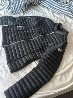 Svart dunjacka från Colmar - Snygg svart dunjacka från Colmar med quiltad design och diskret logga på ärmen. Jackan har dragkedja framtill och en smal, figurnära passform. Perfekt för kyliga dagar och enkel att matcha med olika outfits.