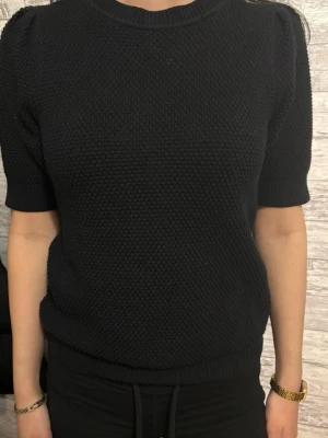 Svart stickad topp med puffärm - Snygg svart stickad topp med korta puffärmar och rund halsringning. Stickad i ett grovt mönster som ger en cool struktur. Perfekt att styla med jeans eller kjol för en trendig look.