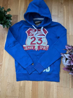 Blå limited edition Superdry hoodie med tryck - Blå hoodie från Superdry med stort vitt och rött tryck på bröstet där det står 'Japan Spirit 23'. Klassisk huva med vita snören och känguruficka framtill. Mjuk insida och ribbade muddar vid ärmar och nederkant. Perfekt för en avslappnad streetwear-look.
