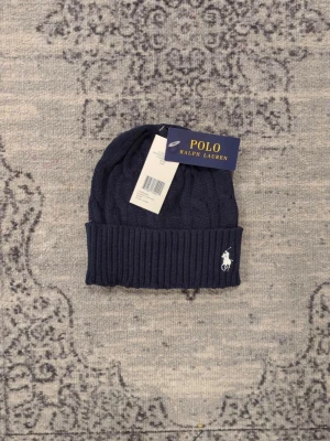 Mörkblå kabelstickad mössa Polo Ralph Lauren - Snygg mörkblå kabelstickad mössa från Polo Ralph Lauren med uppvikt kant och klassisk vit logga broderad framtill. Tillverkad i mjukt material som håller dig varm och stilsäker under kalla dagar.