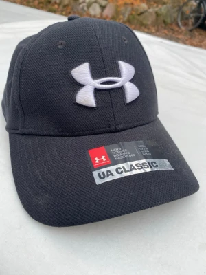 Svart UA Classic keps från Under Armour - Snygg svart keps från Under Armour med klassisk böjd skärm och stort vitt broderat UA-logotyp framtill. Tillverkad i ett ventilerande mesh-liknande material som ger en sportig look. Perfekt för dig som gillar stilrena accessoarer med tydlig logga.