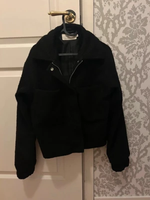 Svart teddyjacka från Nelly - Säljer en svart teddyjacka från Nelly med oversized passform och stora framfickor. Jackan har bred krage, dragkedja och tryckknapp framtill. Perfekt för dig som gillar en trendig och mysig look. Materialet är fluffigt och mjukt.