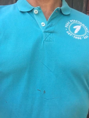 Turkos pikétröja från Peak Performance - Snygg turkos pikétröja från Peak Performance med klassisk krage och två knappar framtill. Tröjan har logga tryckt på bröstet och är tillverkad i ett mjukt bomullsmaterial. Perfekt för en avslappnad och sportig look.
