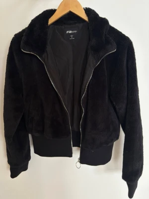 Svart teddyjacka från FB Sister - Mysig svart teddyjacka från FB Sister med mjuk fuskpäls, dragkedja framtill och breda muddar vid ärmslut och midja. Jackan har en hög krage och är helfodrad för extra komfort. Perfekt för dig som vill ha en trendig och varm look.