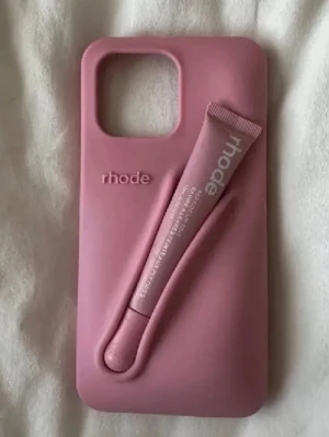 Rosa mobilskal från rhode med hållare - Passar 12,13, 14pro max🫶 Snyggt rosa mobilskal från rhode med smart hållare för läppbalsam eller liknande tub. Skalet är gjort i mjukt silikonmaterial och har en unik design där tuben kan fästas på baksidan. Perfekt för dig som vill ha både stil och funktion i ett.