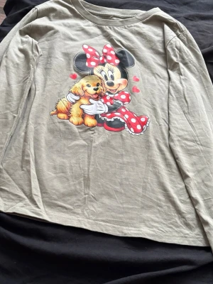 Beige långärmad topp med Minnie Mouse - Söt beige långärmad topp med tryck av Minnie Mouse och en hund på framsidan. Toppen har rund halsringning och är gjord i mjukt bomullsmaterial. Perfekt för dig som gillar Disney och vill ha en bekväm och lekfull stil.