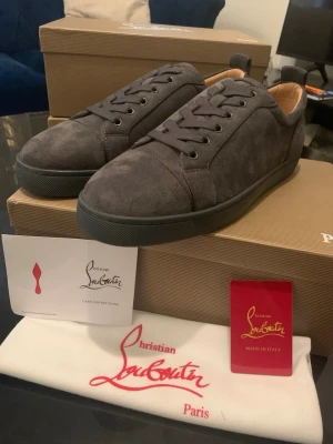 Mörkgrå sneakers från Christian Louboutin - Snygga mörkgrå sneakers i mocka från Christian Louboutin med klassisk röd sula och svarta skosnören. Låg modell med stilren design och diskreta sömmar. Perfekta för dig som vill ha något exklusivt och trendigt i garderoben.