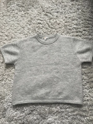 Grå stickad t-shirt Gina Tricot - Snygg grå stickad t-shirt med korta ärmar och rund halsringning. Perfekt för lager-på-lager och enkel att matcha med andra plagg. Använd fåtal gånger. Pris kan diskuteras.