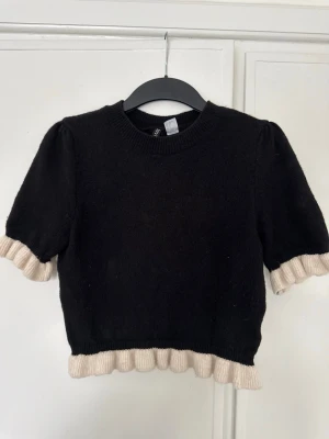 Svart stickad topp med volangkant - Säljer en svart stickad topp med korta puffärmar och beige volangkant vid ärmslut och nederkant. Tröjan har rund halsringning och en croppad passform som ger en trendig look. Perfekt att styla med höga jeans eller kjol.