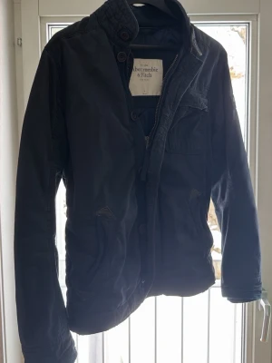 Mörkblå jacka från Abercrombie & Fitch - Snygg mörkblå jacka från Abercrombie & Fitch med hög krage, dragkedja och knappar framtill. Jackan har flera fickor och en patch med logga på ärmen. Perfekt för dig som gillar en stilren och casual look.