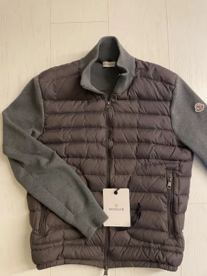 Grå Moncler cardiganjacka - Snygg grå cardiganjacka från Moncler med quiltad front och stickade ärmar. Jackan har dragkedja framtill, två fickor med dragkedja och Moncler-logga på ärmen. Perfekt för lager-på-lager och en clean look.