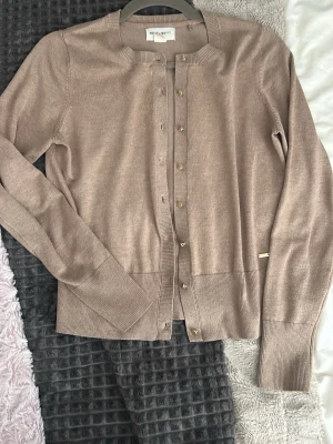 Beige kofta - Snygg beige kofta från Lindex med rund halsringning och guldfärgade knappar. Koftan är ribbstickad vid ärmslut och nederkant, har lång ärm och en liten metallbricka med logga vid midjan. Perfekt att slänga över en topp för en clean look.