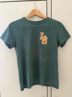 Levi’s T-shirt  - Snygg grön t-shirt med korta ärmar och rund halsringning. På bröstet finns ett gult och vitt LS-tryck som ger en sportig vibe. Perfekt till jeans eller shorts för en avslappnad look.