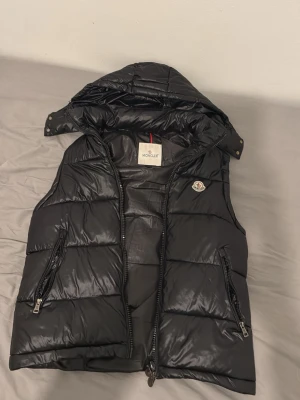 Moncler väst - Snygg svart dunväst från Moncler med avtagbar huva och två dragkedjefickor. Västen har ett glansigt material och klassisk Moncler-logga på bröstet. Perfekt för lager-på-lager och streetwear-vibe. Fin moncler väst i storlek M men passar också S, västen är i fint skick och inga konstigheter med den, kom dm för frågor. Nfc funkar och den går att scanna på badgen