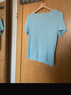 Ljusblå topp från Monki - Supersöt ljusblå topp från Monki med korta ärmar och vågiga kanter. Tillverkad i ett stretchigt, ribbat material som ger en skön och avslappnad look. 