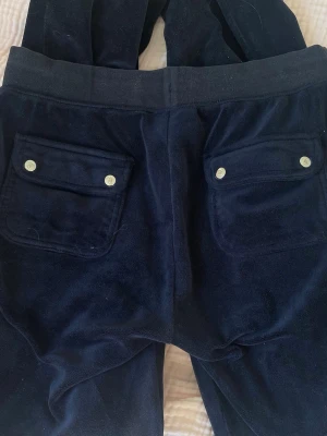 Mörkblå velourbyxor Juicy Couture - Mörkblå mjukisbyxor i velour från Juicy Couture med bred resår i midjan och dragsko med gulddetaljer. Byxorna har stora fickor både fram och bak, där bakfickorna har lock och tryckknappar. 