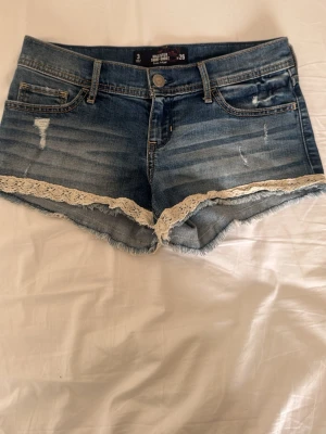 Blå jeansshorts med spetskant - Snygga blå jeansshorts från Hollister med slitna detaljer och vit spetskant längs bensluten. Klassisk femficksmodell med knapp och dragkedja framtill. Perfekta för sommaren!💗I stretchigt material.