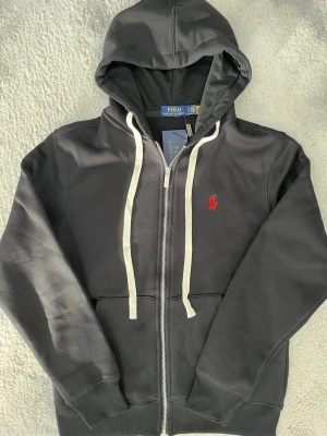 Svart hoodie från Polo Ralph Lauren - Svart hoodie från Polo Ralph Lauren med vit dragkedja och snörning. Klassisk röd logga broderad på bröstet. Tillverkad i mjuk bomull och har kängurufickor framtill. Perfekt för en chill och stilren look. Väldigt liten i storlek, storlek M men passar storlek XS