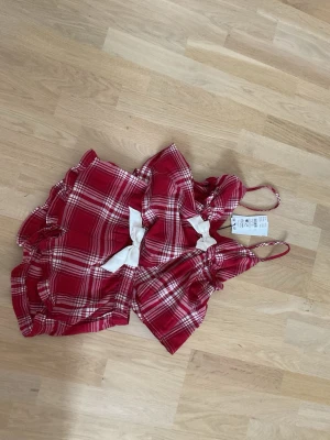 Röd rutig pyjamas 2 delad - Supersöt röd och vit rutig pyjamas med tunna axelband och dekorativ vit rosett framtill. Shorts och line. Tillverkad i mjukt tyg, perfekt för dig som gillar en lekfull och trendig stil. Volangdetaljer längs kanterna ger en extra feminin touch.