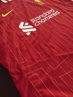 Liverpool matchtröja Nike röd - Liverpool FC matchtröja i rött med gula detaljer och tunna gula ränder. Tröjan har klubbmärke och Nike-logga på bröstet samt sponsortryck från Standard Chartered. Klassisk krage med vita och gula inslag. Perfekt för fotboll och sport. Är storlek XXL men känns som L/XL