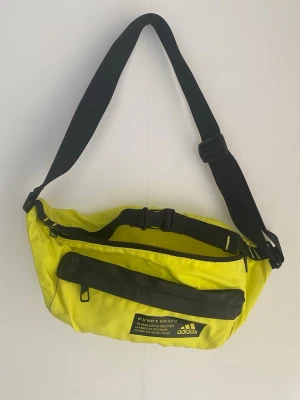 Gul magväska från Adidas - Cool gul magväska från Adidas med svart justerbart axelband och svart dragkedja. Väskan har en stor ytterficka och Adidas-logga framtill. Perfekt för att bära det viktigaste och ge din outfit en sportig touch.