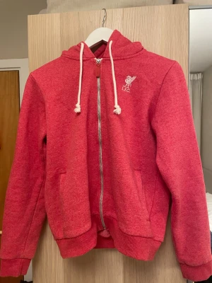 Röd LFC zip hoodie med huva - Röd hoodie med dragkedja från Liverpool FC. Broderad L.F.C.-logga på bröstet och vita dragsnören i huvan. Mjuk bomullsblandning, ribbade muddar och två fickor framtill. Perfekt för dig som vill visa ditt lagstöd på ett stilrent sätt.