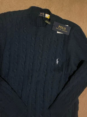 Stickad tröja  - Mörkblå kabelstickad tröja med rund halsringning och klassisk broderad logga på bröstet. Tröjan har långa ärmar och ribbade muddar. Perfekt för dig som gillar stilrena och tidlösa plagg.