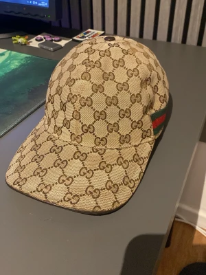 Beige GG-mönstrad keps från Gucci - Säljer en beige keps från Gucci med det klassiska GG-monogrammet över hela kepsen. På sidan finns den ikoniska gröna och röda randen. Kepsen har böjd skärm och justerbar rem bak. Materialet är canvas med exklusiv känsla.