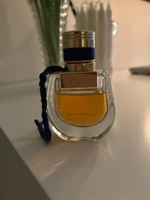 Chloé Nomade Eau de Parfum - Elegant parfym från Chloé i en genomskinlig glasflaska med gulddetaljer och mörkblått band runt halsen. Flaskan har en rundad form och innehållet är bärnstensfärgat. Perfekt för dig som gillar lyxiga och feminina dofter. Den är 30 ml men finns 25 ml kvar. Tyvärr ingen kartong!  - Titta gärna på mina andra annonser!! 