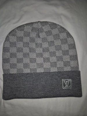 Grå Louis Vuitton mössa med rutmönster - Snygg grå mössa från Louis Vuitton med klassiskt rutmönster och vikt kant. LV-logga broderad framtill på kanten. Tillverkad i mjukt stickat material som håller dig varm och stilren under kalla dagar.