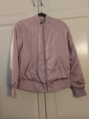 Ljusrosa bomberjacka från H&M - Snygg ljusrosa bomberjacka från H&M med glansigt tyg och klassisk ribbad mudd vid ärmslut och midja. Jackan har dragkedja framtill och två snedställda fickor med knapp. Perfekt för dig som vill ha en stilren och trendig look.