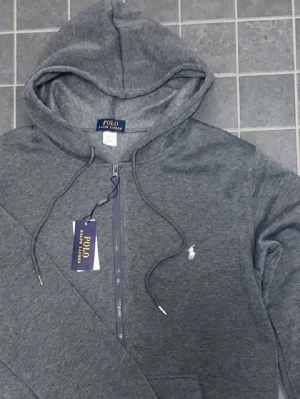 Grå hoodie från Polo Ralph Lauren - Pris kan diskuteras. Mörkgrå hoodie med dragkedja från Polo Ralph Lauren. Klassisk design med huva, dragsnören och vit broderad logga på bröstet. Tillverkad i mjukt material som är skönt att bära. Perfekt för en avslappnad och stilren look.