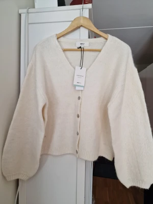 Creme vit kofta från Gina Tricot - Mysig kofta från Gina i stl S med v-ringning och stora, vida ärmar. Koftan har en croppad passform och stängs med fem knappar framtill. Tillverkad i ett mjukt, stickat material som känns lyxigt och trendigt.