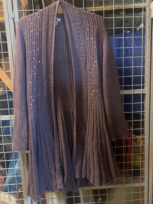 Lila lång kofta med paljetter - Säljer en lila lång stickad kofta med öppen front och dekorativa paljetter längs framstyckena. Koftan har ett fint fall och ribbade detaljer som ger extra struktur. Perfekt för dig som vill ha något både snyggt och bekvämt.