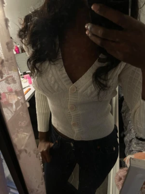 Vit ribbstickad kofta med knappar - Snygg vit ribbstickad kofta med kabelstickat mönster och stora knappar framtill. Koftan har djup v-ringning och sitter tajt i midjan, perfekt att styla med jeans. Långa ärmar och mjukt material som känns skönt mot huden.