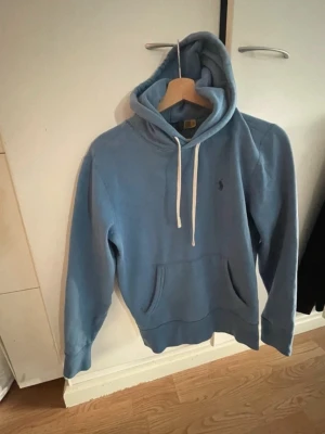 Blå hoodie från Polo Ralph Lauren - Säljer en ljusblå hoodie från Polo Ralph Lauren med klassisk broderad logga på bröstet. Hoodien har huva med vita snören och en stor magficka framtill. Tillverkad i mjukt bomullsmaterial och har ribbade muddar vid ärmar och nederkant i storlek xs 
