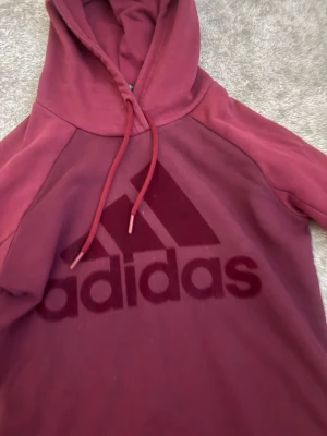 Vinröd hoodie från Adidas - Snygg vinröd hoodie från Adidas med stor logga på bröstet. Hoodien har långa ärmar, dragsko i huvan och är tillverkad i mjukt material som passar perfekt för en avslappnad stil. Klassisk passform och sportig look.
