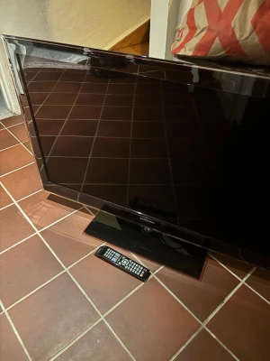 Samsung TV med fjärrkontroll - Säljer en svart platt-TV från Samsung med tillhörande fjärrkontroll. TV:n har en stilren design med bred ram och står på en rektangulär fot. Perfekt för filmkvällar eller gaming. Passar bra i vardagsrummet eller sovrummet.