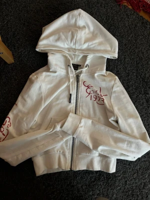 Vit Ed Hardy hoodie med färgglad print - Säljer en vit croppad hoodie från Ed Hardy med dragkedja, huva och ribbade muddar. Framsidan har röd text och årtal, medan baksidan pryds av en stor färgglad fågel och blommor samt Ed Hardy-loggan. Tryck även på ena ärmen. Perfekt för dig som gillar statement-plagg!